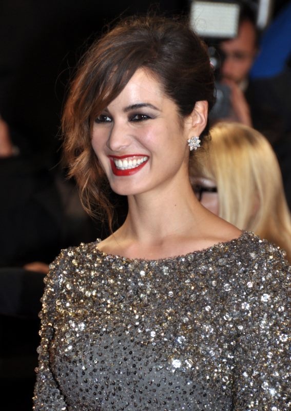 A smiling woman with dark hair pinned up, red lipstick and a sparkling silver sequined top, with a photographer and other guests blurred in the dark background.