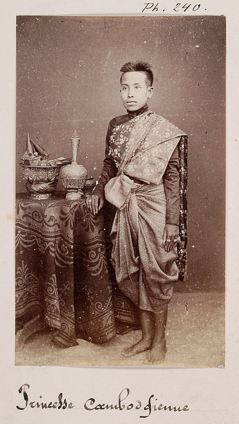 A woman standing beside a draped table with a vase, wearing a patterned shawl over one shoulder and a sampot pulled up between the legs.