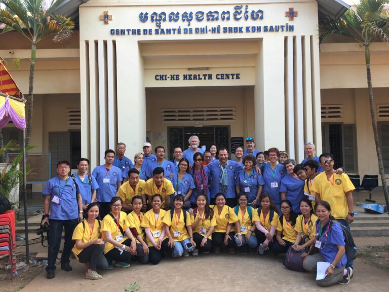 A large team in blue scrubs and yellow shirts posed in front of a health centre with its name in Khmer and French.