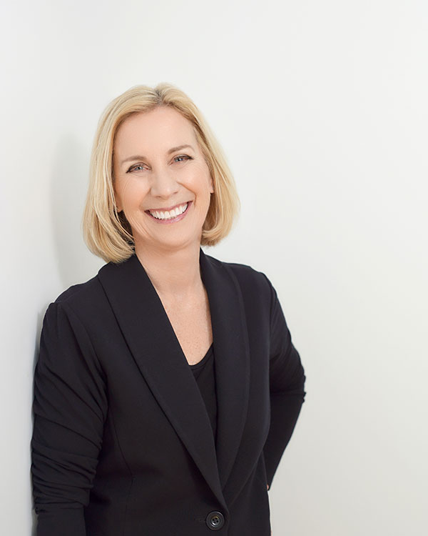 A smiling woman with shoulder-length blond hair in a black jacket against a white wall.