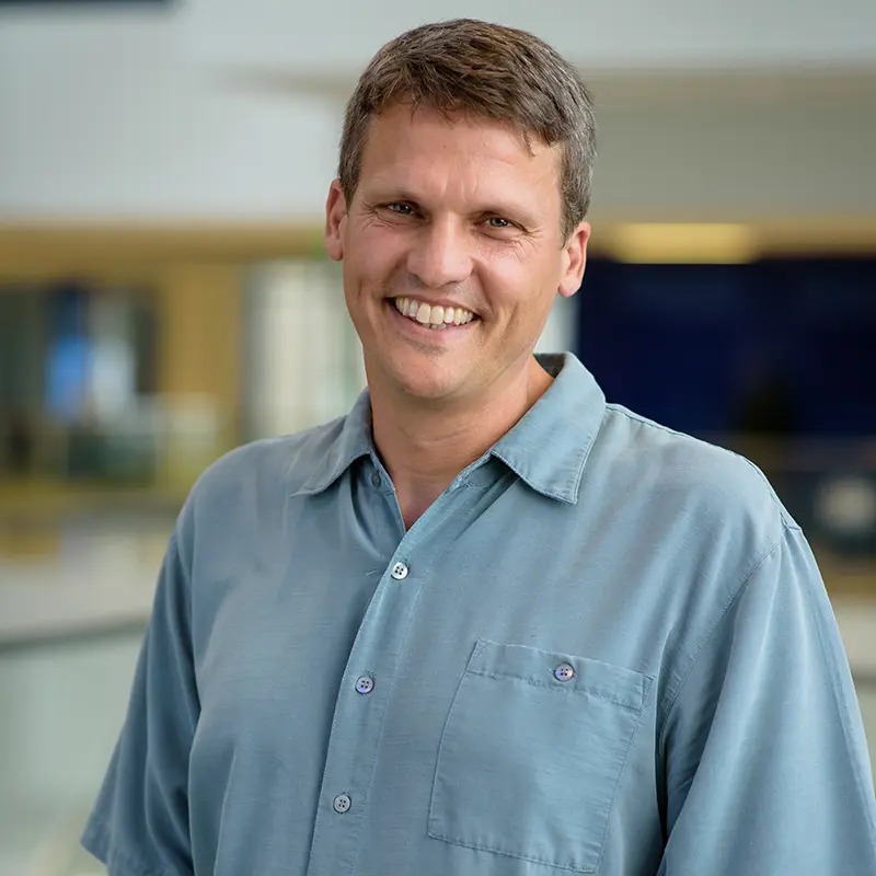A smiling man in a blue short-sleeved shirt standing in a bright building.