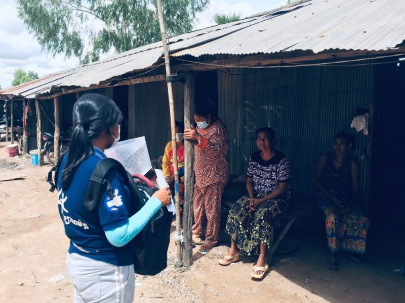 A worker in a blue Friends International shirt with a backpack talks with adults sitting outside tin-roofed houses.