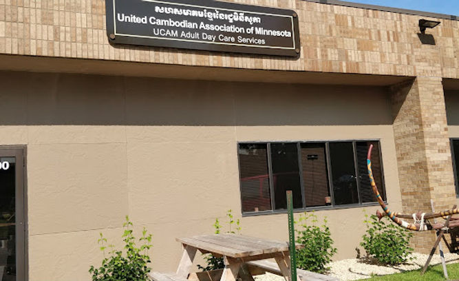 A tan stone building front with a dark sign in Khmer and English and a picnic table outside.