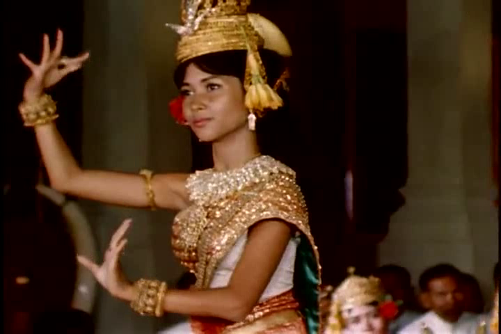 A dancer in a gold crown and embroidered costume raises one hand with fingers curved back.