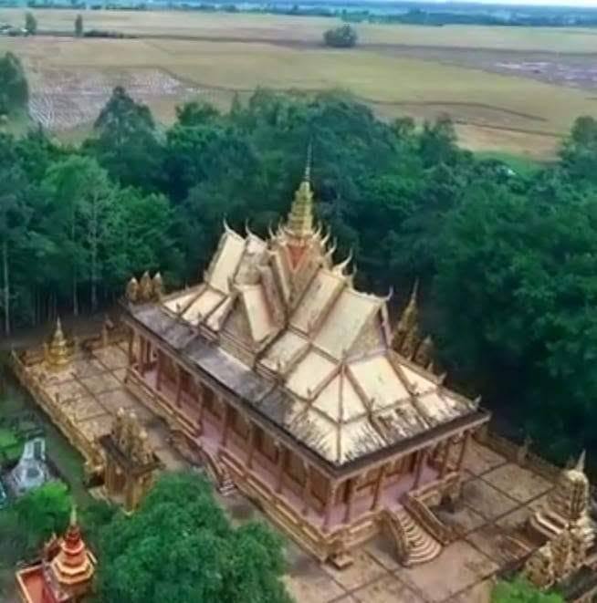 An aerial view of a Khmer temple hall with a tiered roof among trees, with rice fields behind.