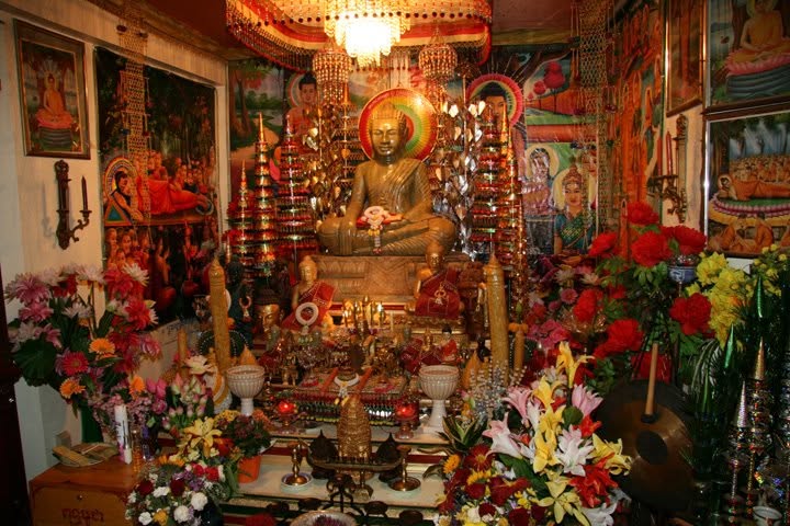 A seated Buddha on a crowded altar with flowers, candles, banners and paintings.