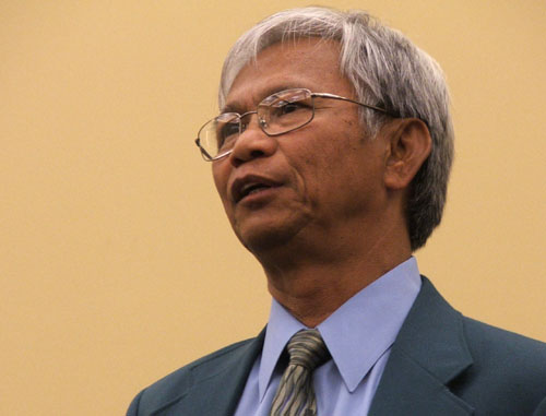 A grey-haired man with glasses in a dark suit and tie, looking up as he speaks.