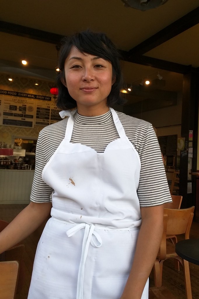 A woman in a white apron and striped shirt stands at the entrance of a cafe.