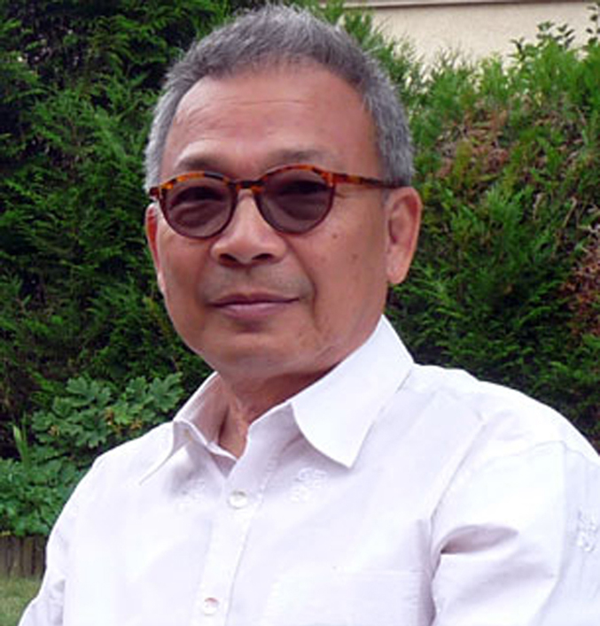 Portrait of an older man with grey hair, tinted round glasses and a white shirt, in front of a green hedge.