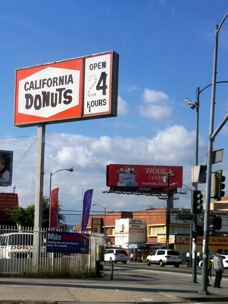 A tall red and white roadside sign reading California Donuts, Open 24 Hours, above a street corner under a blue sky.