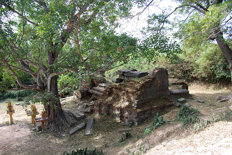 A low, broken brick and stone structure under large trees, with fallen blocks around it.