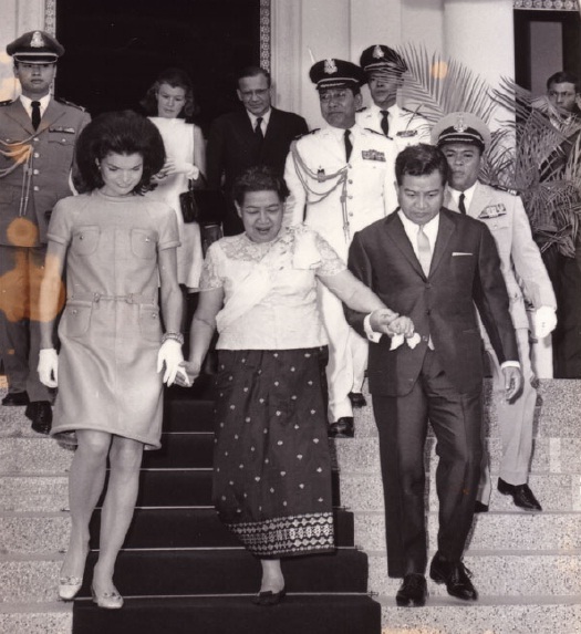Three people walk down palace steps, the queen in the centre in a white blouse and patterned sampot, officials behind.