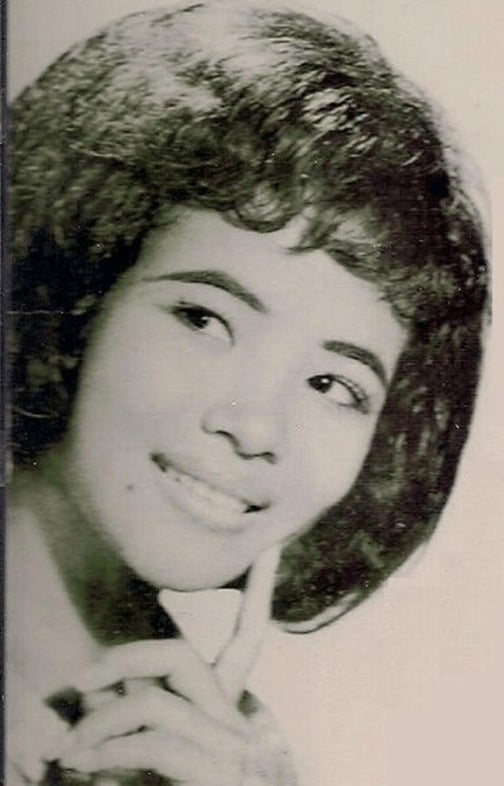 A black-and-white portrait of a smiling young woman with full, curled dark hair, one finger resting against her chin.
