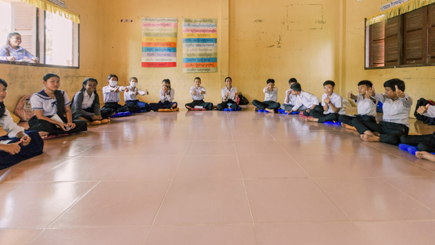 School students in white shirts sit in a circle along the walls of a large yellow room with posters on the wall.