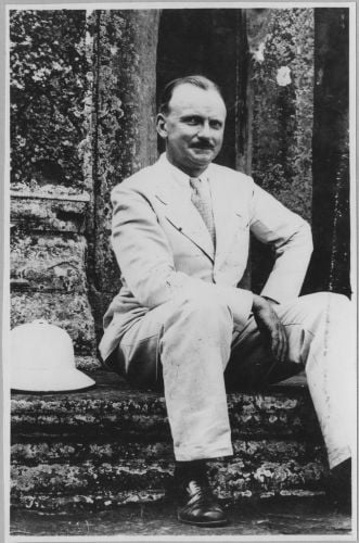 A black and white photograph of a man in a light suit sitting on stone steps, a pith helmet beside him.