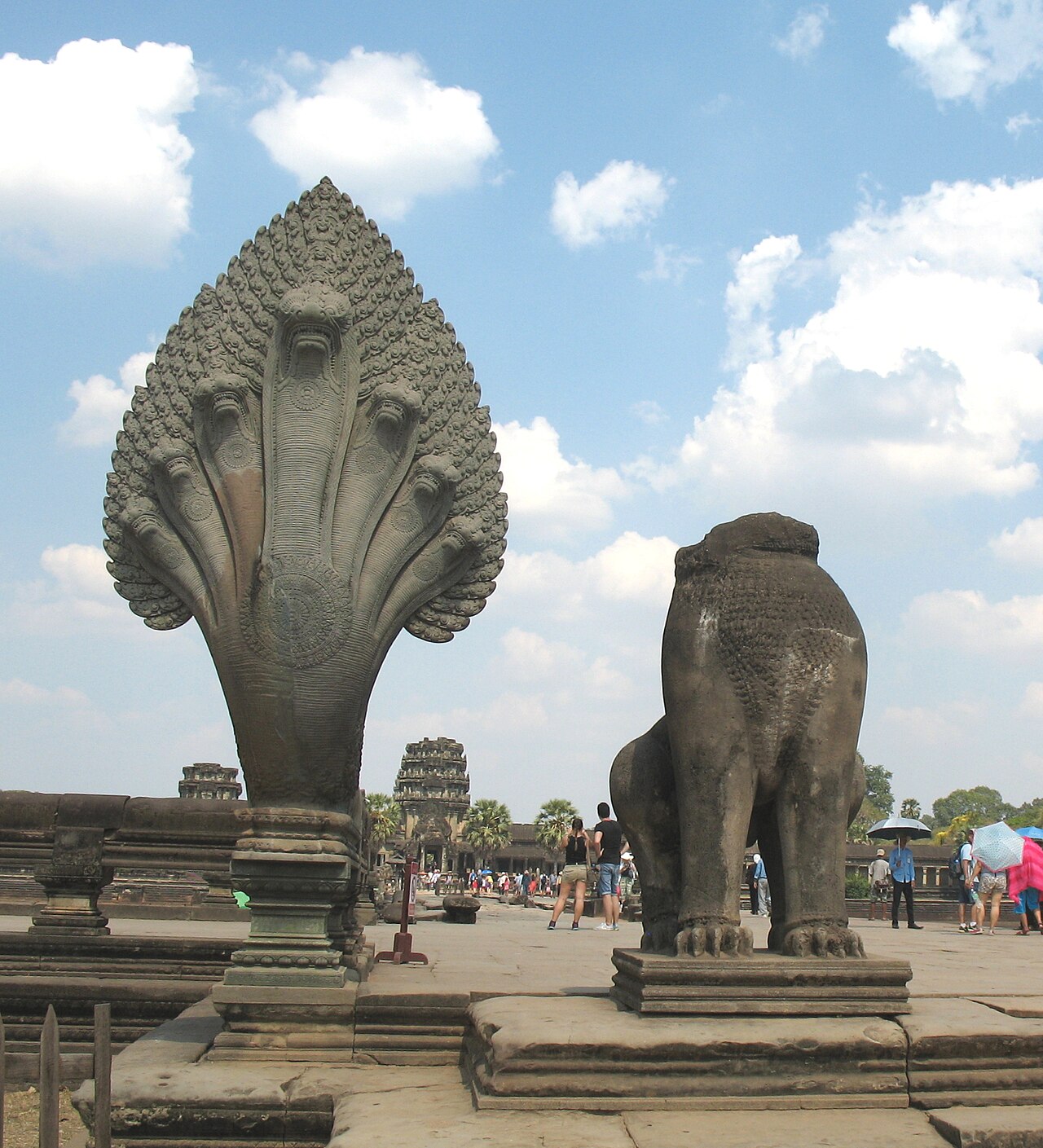 A seven-headed stone naga beside a stone lion seen from behind on a sandstone terrace, with visitors and temple towers in the distance.