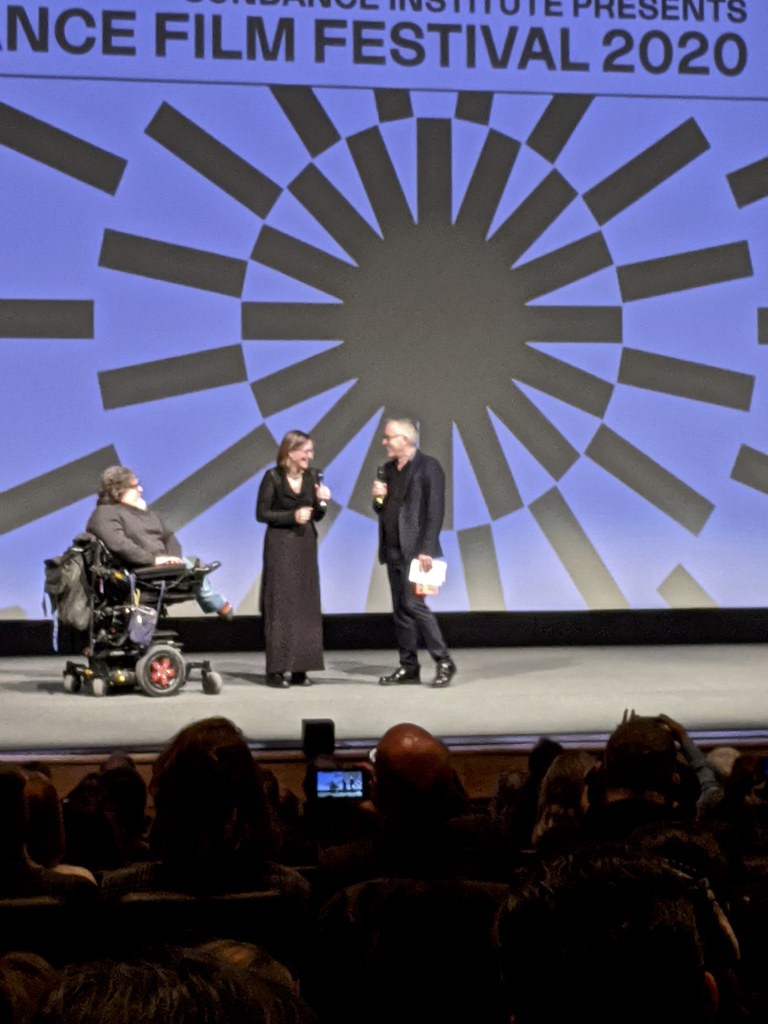 Three people on a theater stage in front of a large blue Sundance Film Festival 2020 backdrop, with the audience in the foreground.