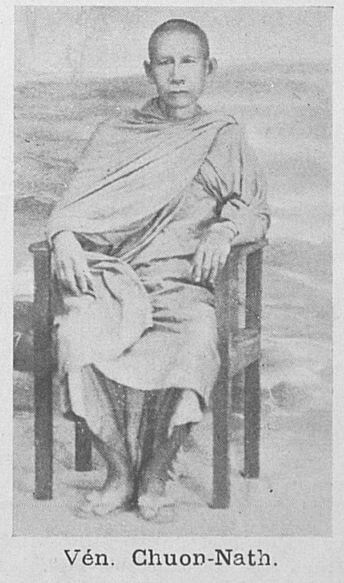 A black-and-white photograph of a monk with a shaved head, wearing robes draped over one shoulder, seated on a wooden chair with his hands resting on his knees. A printed caption below reads "Vén. Chuon-Nath."