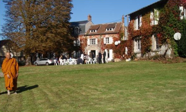 A large ivy-covered stone house across a lawn, with a monk in robes in the foreground and guests at tables.