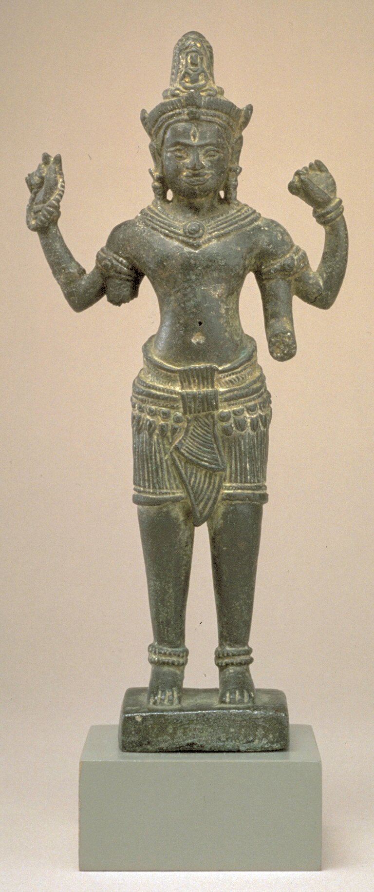 A dark bronze figure standing with four arms and a tall headdress, on a square base.