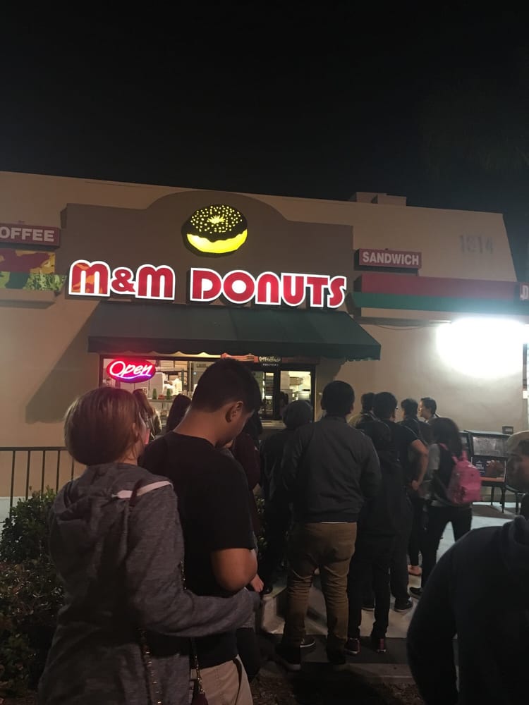 Lit shop sign reading M&M Donuts with a glowing donut above it, and a line of people outside at night.
