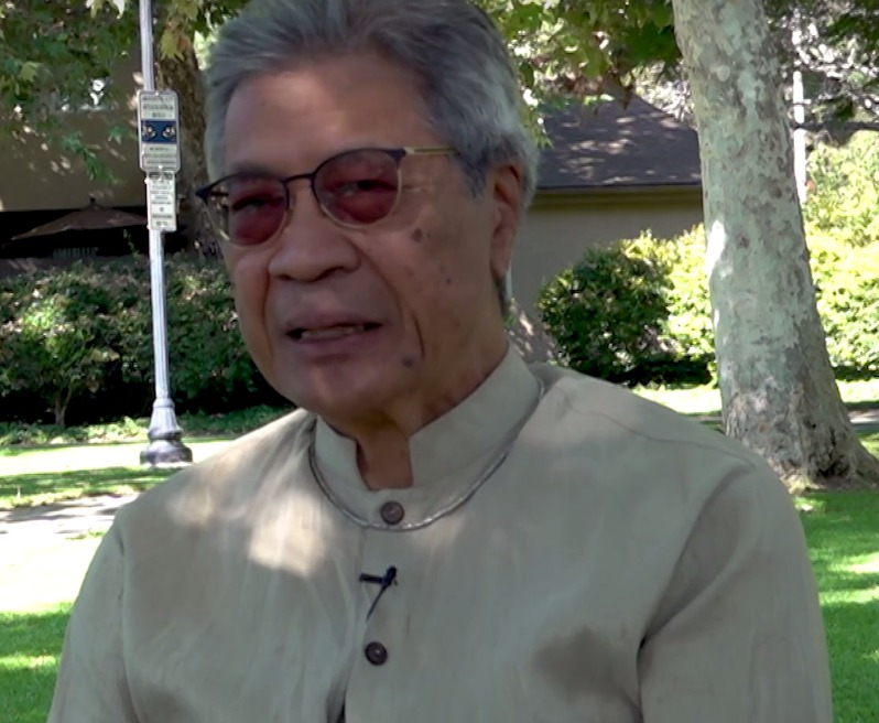 Chinary Ung, with gray hair and tinted glasses, in a beige shirt with a standing collar, speaking outdoors under trees.