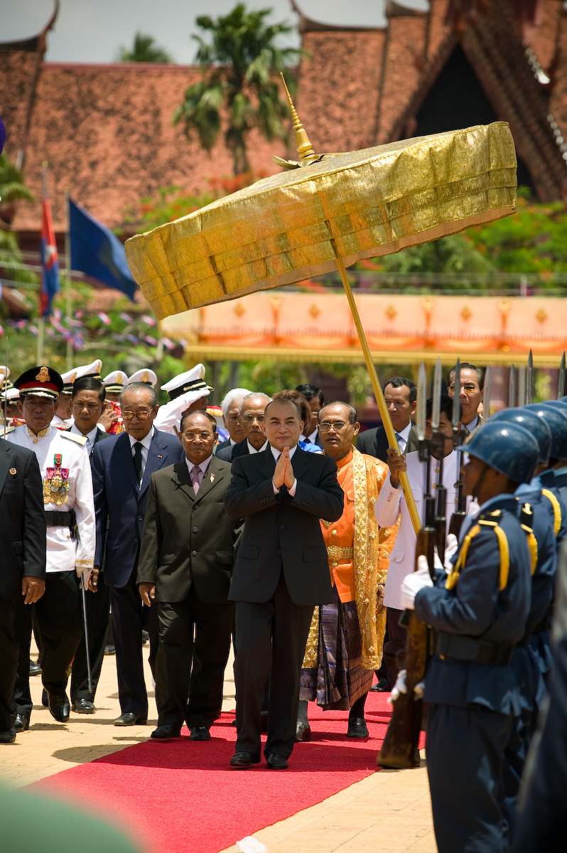 The king in a dark suit walks on a red carpet with his palms pressed together, under a gold ceremonial parasol, between uniformed guards and officials.