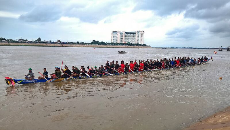 A long racing boat packed with rowers in mixed shirts on a wide brown river, with a tall hotel on the far bank.