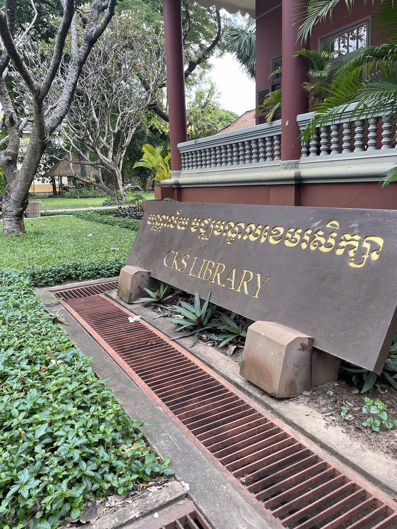 A stone sign reading CKS Library in Khmer and English beside a path, with a wooden building and trees behind.