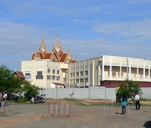 A long cream-colored building with rows of windows and a long wing to the right, topped by a red and gold roof with three tall golden spires, stands behind a metal fence across a wide dirt lot with crowd barriers, a few people and parked motorbikes, under a blue sky.