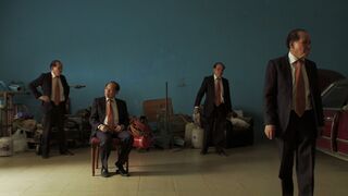 Four men in dark suits and orange ties in a bare blue-walled room cluttered with old furniture, one seated on a chair.