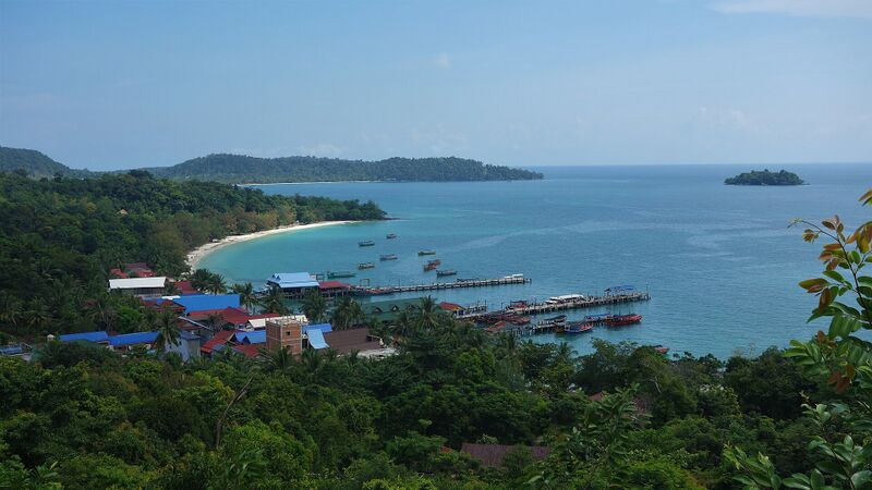 A view from a hillside over a small pier and bay of turquoise water, with forested islands on the horizon.