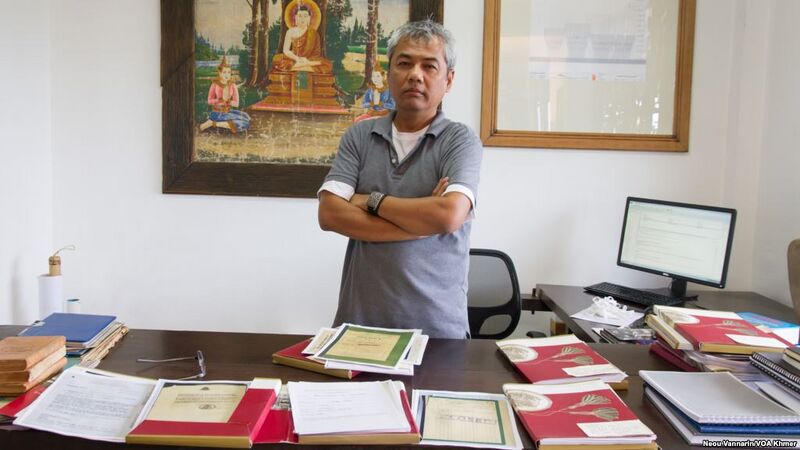 A man with gray hair stands with folded arms behind a desk covered with open documents.