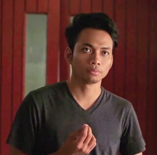 A young man in a grey T-shirt talks to the camera in front of a red wooden wall.