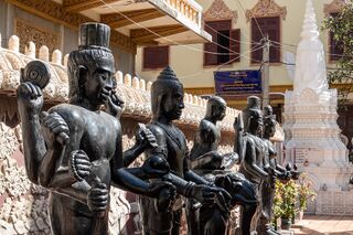 Several dark standing statues of figures lined up in front of a carved white wall and a small stupa.