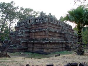 A stepped pyramid of reddish stone, seen from one corner, rises in three tiers to a ruined gallery with small windows on top. Stone lions guard a stairway on the left, with tall trees behind and a fan palm on the right.