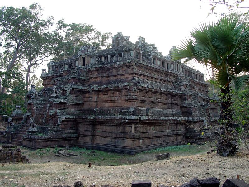 A stepped pyramid of reddish stone, seen from one corner, rises in three tiers to a ruined gallery with small windows on top. Stone lions guard a stairway on the left, with tall trees behind and a fan palm on the right.