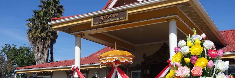 Temple porch with a sign in Khmer and English, a gold ceremonial parasol and large artificial flower arrangements, under a blue sky.