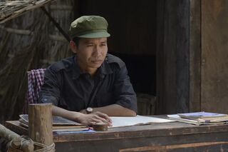 A man in a dark shirt and green cap sits writing at a wooden desk in a bamboo hut.