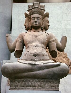 A sandstone statue of a seated god with four crowned heads, three of them facing the viewer, sits cross-legged on a carved base. Its four arms are broken off below the elbow, and cracks run across the chest, against a pale gray museum wall.