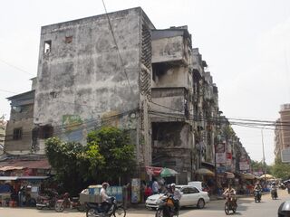 A weathered four-storey concrete apartment block with shops and motorbikes on the street in front.