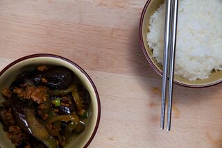 A bowl of dark stir-fried eggplant and minced pork next to a bowl of white rice and chopsticks on a wooden board.