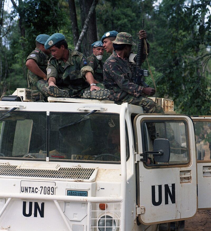 Soldiers in camouflage and blue berets sit on top of a white vehicle marked UN and UNTAC-7089, with forest behind.