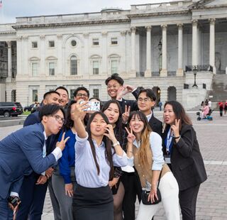 A group of young people in business clothes take a selfie in front of the white columned Capitol building.