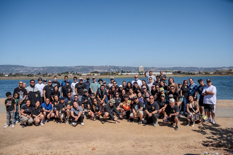 A large group of adults and some children posing together on a sandy beach under a clear blue sky.