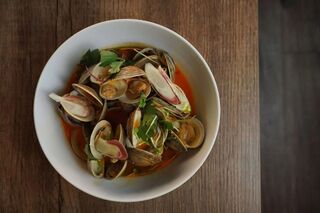 White bowl of clams in a light broth with sliced chilli and herbs on a dark wooden table.