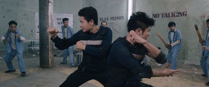 Two men in black fight in a prison hall while inmates in blue watch from the sides.