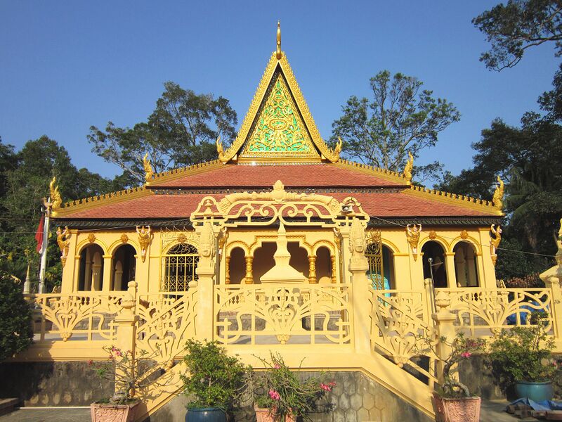 The yellow facade of a Khmer temple hall with arched openings, a red tiered roof and a gilded gable, among trees.