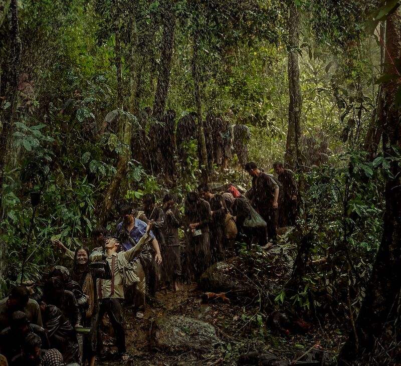 Dozens of people in worn clothes walk in a line along a muddy forest trail in heavy rain, some carrying bundles.