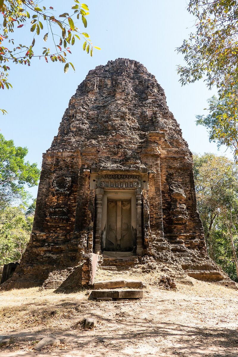 A weathered brick temple tower with a doorway and a short stair, standing among trees under a blue sky.
